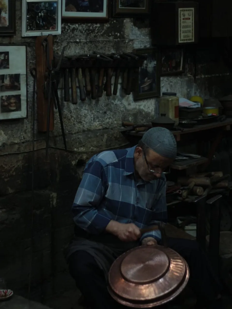 Artesano trabajando una bandeja de cobre con martillo en un taller, con juegos de cinceles colgados en la pared